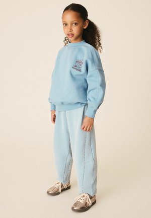 Ung pige med krøllet hår iført en lyseblå sweatshirt med et husdesign, blåstribede bukser og metalliske gyldne sneakers, stående.