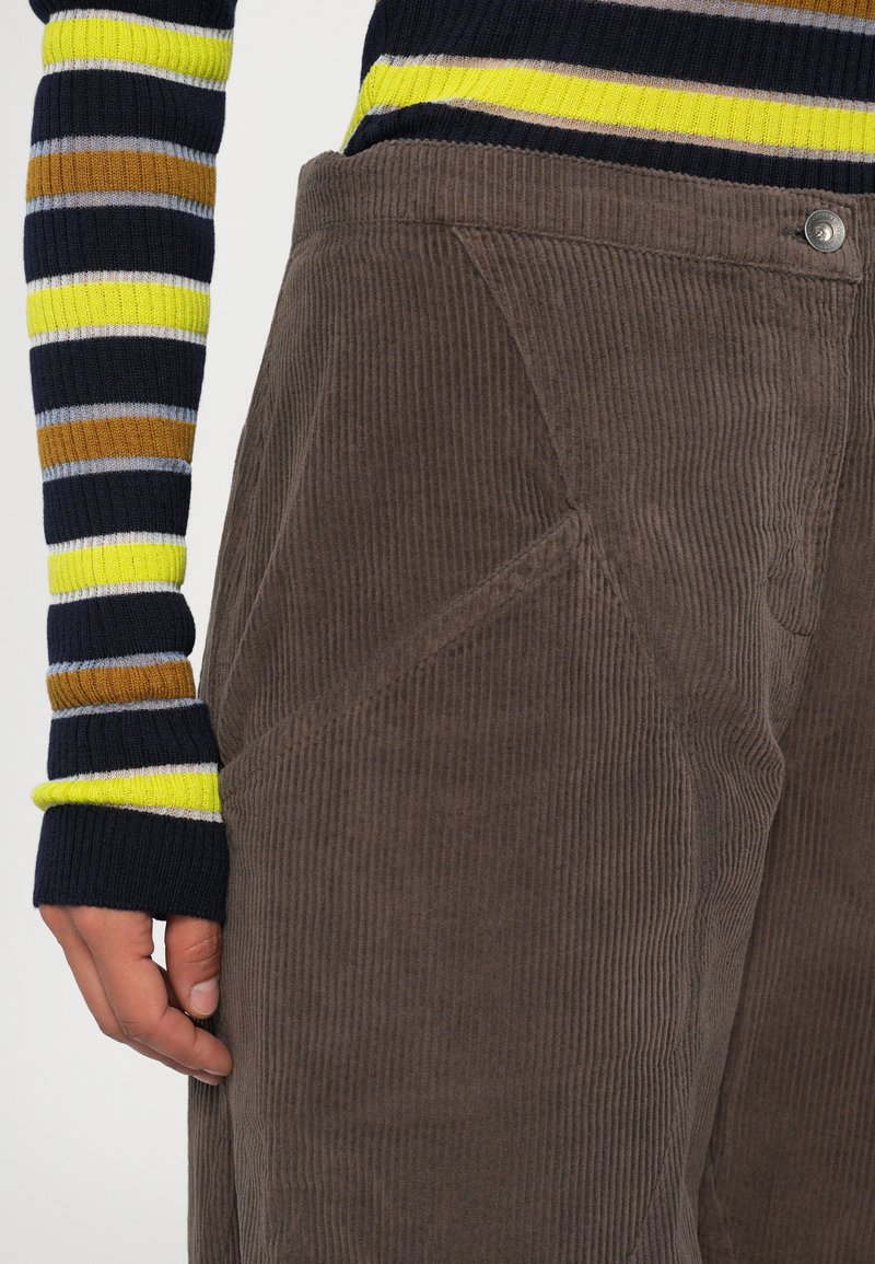 Mano vicino all'anca che indossa pantaloni in velluto a coste marroni e un maglione a maniche lunghe a righe multicolori con righe blu navy, giallo, marrone e azzurro chiaro.