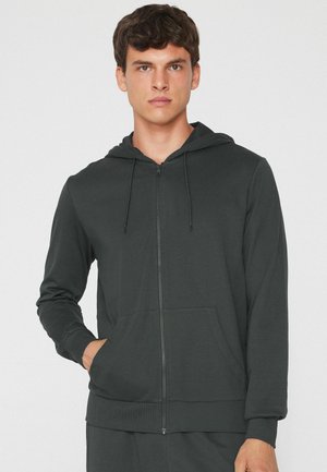 Junger Mann trägt einen dunklen Kapuzenpullover mit Reißverschluss, Fronttaschen und Kapuze, steht vor hellem, einfarbigem Hintergrund.