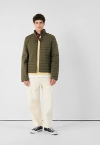 Joven vestido con una chaqueta acolchada verde oliva, pantalones beige, zapatillas negras y un pañuelo rojo de cuello, de pie frente a un fondo blanco liso.