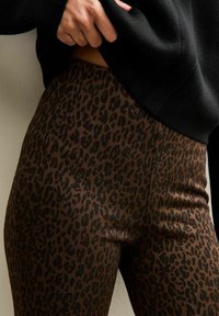 Gros plan sur une main soulevant un pull noir, révélant un legging marron taille haute avec un motif léopard noir.