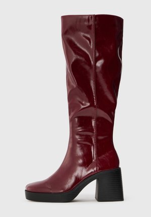 Botas burdeos hasta la rodilla con un acabado brillante. Punta cuadrada, tacón grueso y un diseño elegante con pliegues sutiles a lo largo del caña.