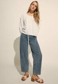 Hvid langærmet bluse med snore ved halsudskæringen, parret med løse blå denim jeans og beige remsandaler med kilehæl i træ.
