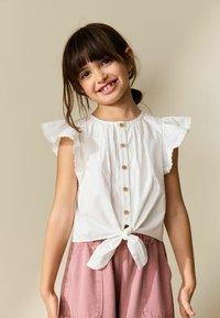 Balts kokvilnas tops ar plandošām piedurknēm, pogu aizdari priekšpusē un sasietu apakšu, kombinēts ar rozā auduma šortiem. Gluda tekstūra, ikdienas dizains.