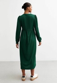 Grön, midi-lång klänning med långa ärmar och volang i midjan. Texturerad tyg och figursydd överdel, matchad med vita skor med svarta detaljer.