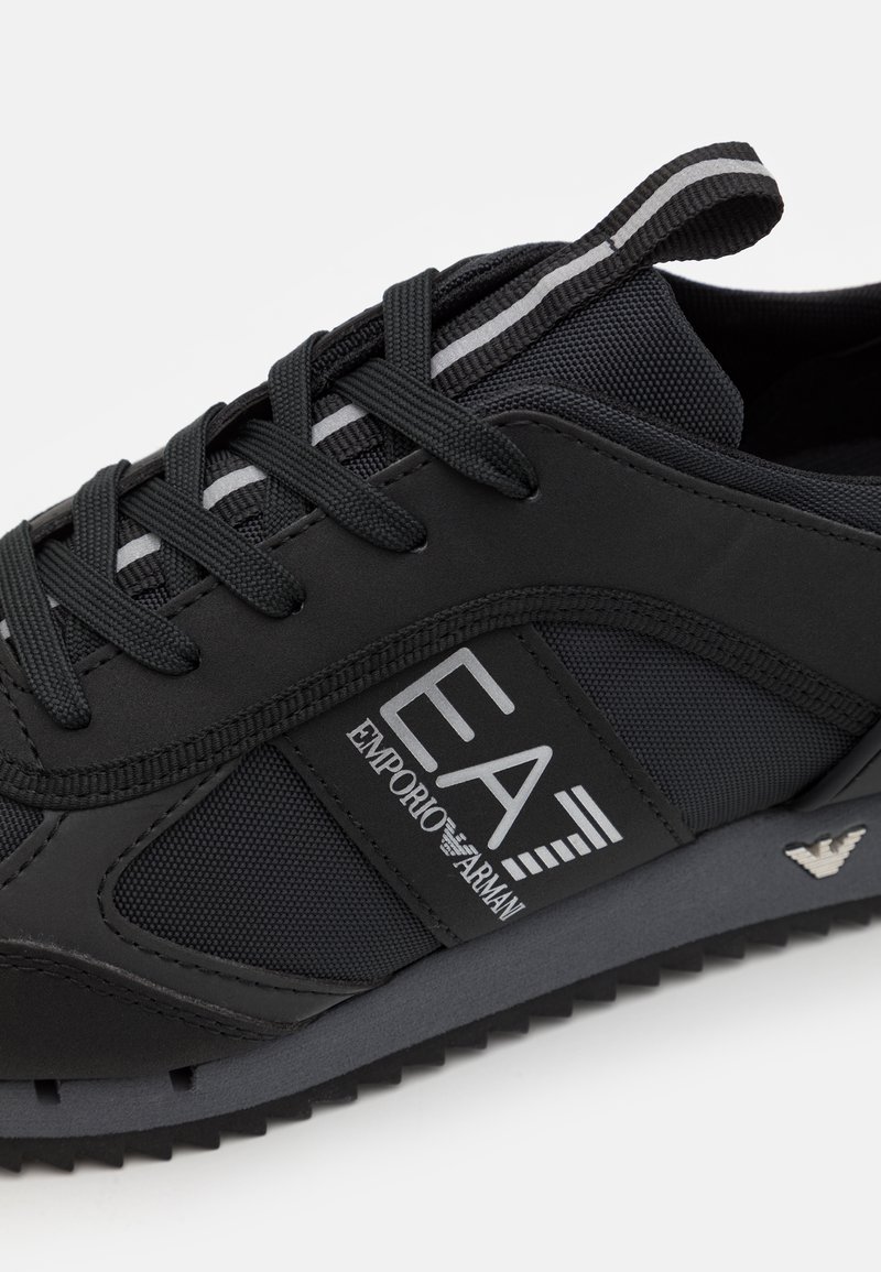 Scarpa sportiva nera con tomaia in mesh e sintetico, lacci e logo in rilievo. Suola distintiva con motivo di trazione e accenti riflettenti.