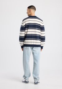 Marineblauw en crème gestreepte gebreide trui met een ronde hals, gecombineerd met losse lichtblauwe jeans en witte sneakers met donkere accenten.