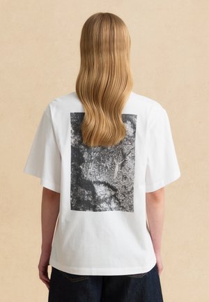 Personne avec de longs cheveux blonds ondulés portant un t-shirt blanc avec un motif abstrait noir et blanc dans le dos.