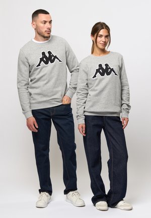 Grijze katoenen sweatshirts met een zwart grafisch logo op de voorkant, gecombineerd met donkerdenim jeans en witte sneakers. Unisex ontwerp.