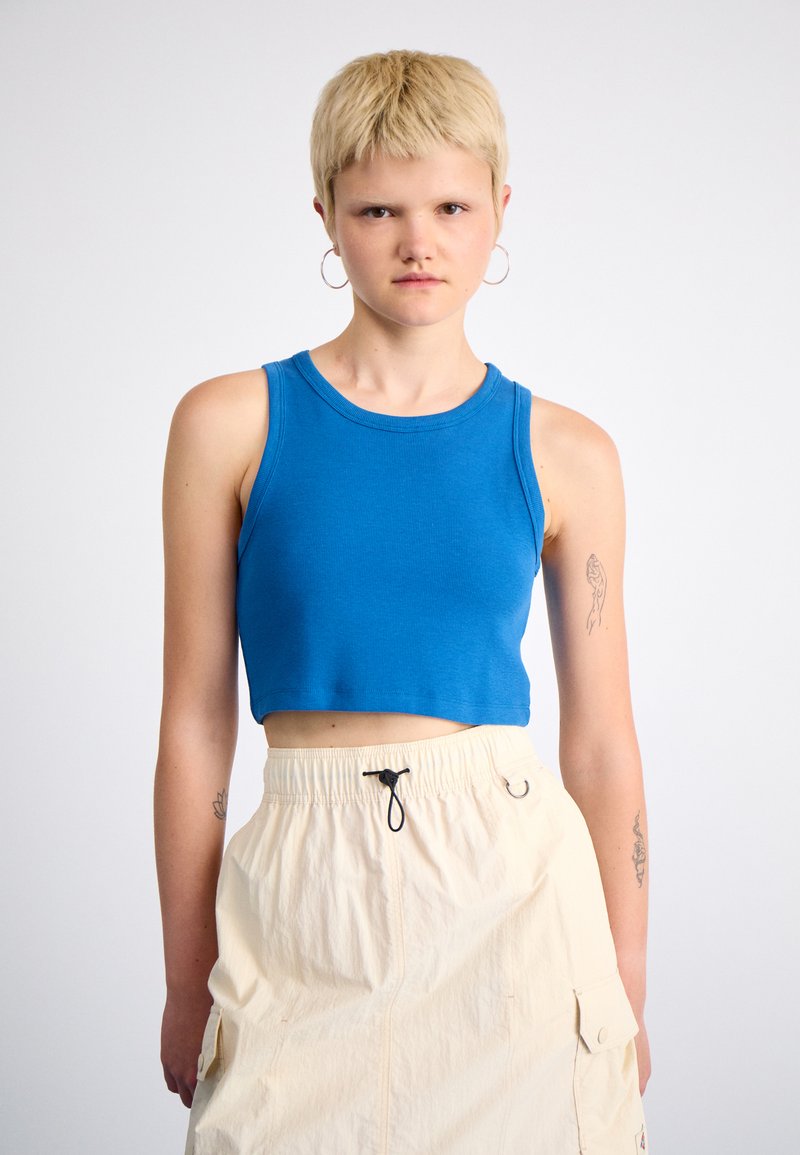 Modrý riasený crop top s okrúhlym výstrihom, skombinovaný s béžovou cargo sukňou na šnúrku, ktorá má bočné vrecká. Modelka má krátke vlasy.