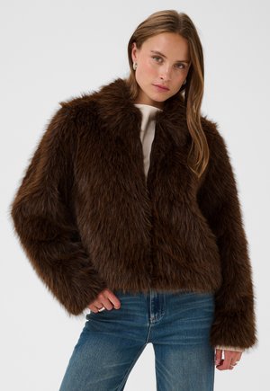 Jeune femme portant une veste en fausse fourrure marron foncé, un haut clair et un jean bleu, debout devant un fond uni.
