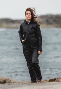 Giacca nera impermeabile con zip frontale, cappuccio e polsini elasticizzati, abbinata a pantaloni coordinati. Texture liscia, design elegante e minimal con poca o nessuna presenza di logo.