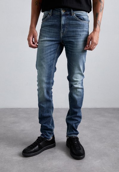Slim-fit siniset farkut, joissa on haalistuneet reidet ja kulutuksia. Yhdistetty mustiin tennareihin. Ompeluyksityiskohtia lähellä taskuja.