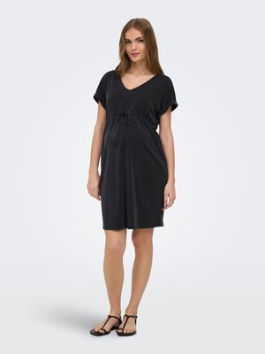 Mujer embarazada de pie, vistiendo un vestido premamá negro hasta la rodilla con mangas cortas y sandalias negras, sobre un fondo liso.