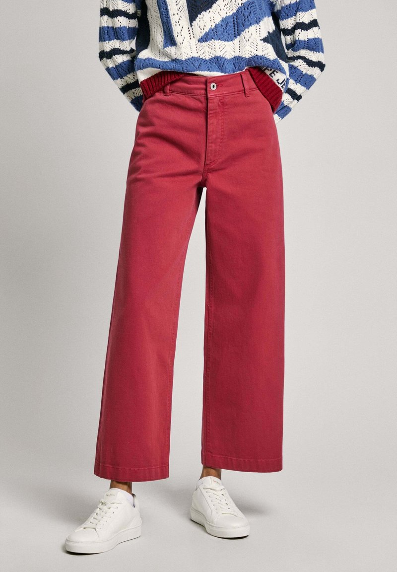 Personne portant un pantalon rouge taille haute à jambes larges, des baskets blanches et un pull en maille rayé bleu, blanc et marine, les mains dans les poches.