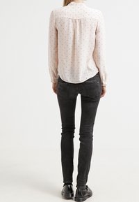 Blouse longue à manches longues rose clair avec des pois noirs ; ourlet incurvé ; associée à un jean noir ajusté et des chaussures noires.