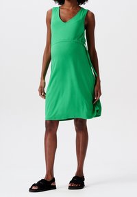 Vestido verde sin mangas con escote en V, fabricado en una tela suave. Detalle de lazo en el costado, de longitud media, combinado con sandalias negras. Textura suave.