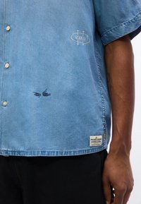 Μπλε κοντομάνικο πουκάμισο με κουμπιά, με γραφικό "RODEO" και μικρή σχεδίαση ματιών, φορεμένο από άτομο με σκουρόχρωμο δέρμα και μαύρο παντελόνι.