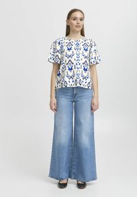 Top corto bianco con motivo astratto blu, abbinato a jeans a gamba larga azzurri. Il modello è in piedi su uno sfondo neutro, indossando scarpe nere.
