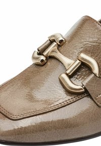 Beige lädersko med en blank yta, rundad form och ett dekorativt spänne i guldton med ribbad design.
