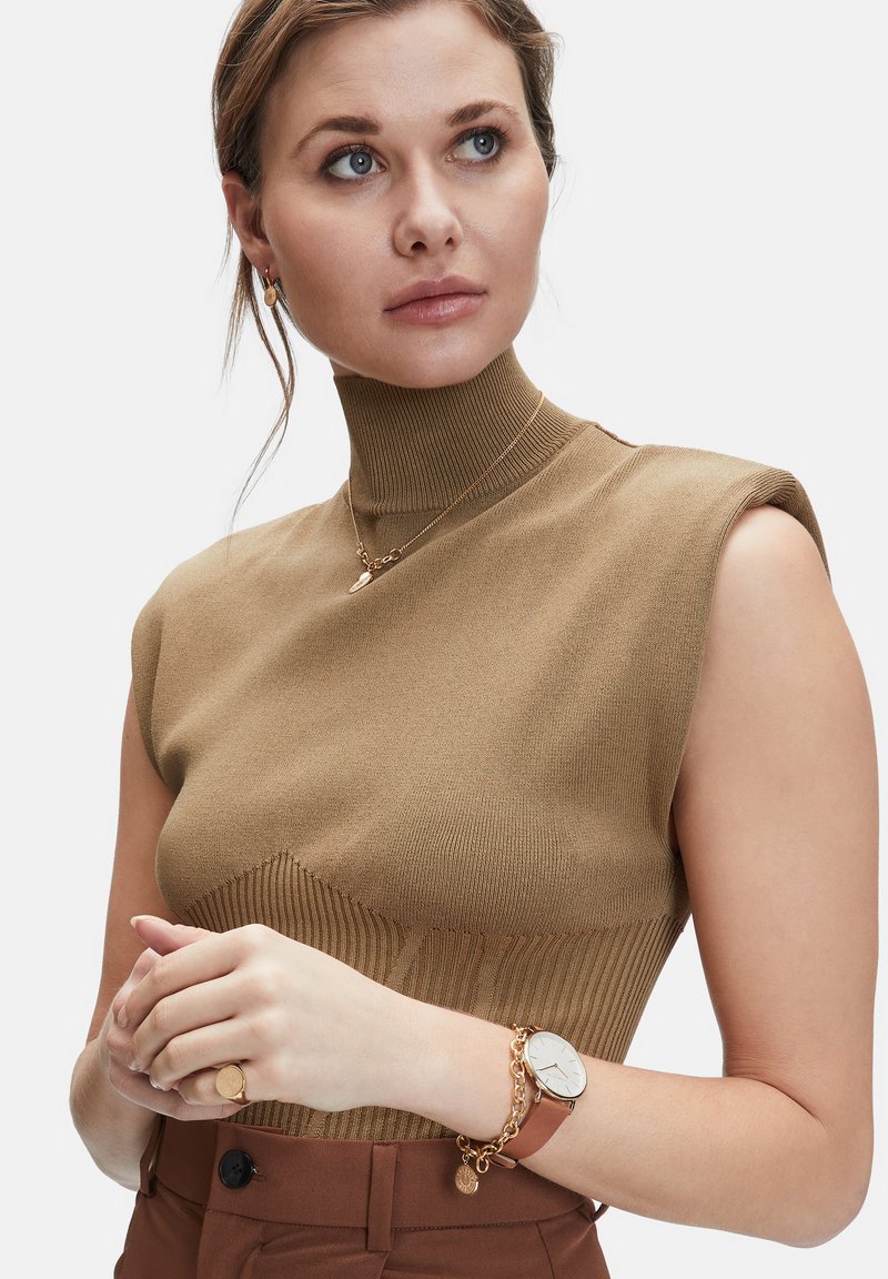 Brun ribbet strikbluse med høj hals; modellen tilbehør med guld smykker og brun ur, præsenteret mod en hvid baggrund.