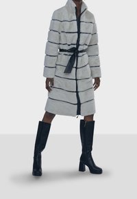 Kabát z umělé kožešiny v krémové barvě s černými pruhy, s celoprsým zipem a odpovídajícím páskem v pase. Kombinováno s černými kozačkami až k kolenům.