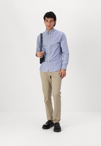 Camisa de botones azul claro, pantalones beige y zapatos negros. La camisa tiene un cuello y una textura sutil, con un pequeño logo en el pecho.