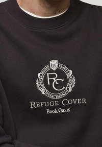 Sudadera negra con un logo circular bordado, texto que dice "Refuge Cover" y "Book Oasis", con detalles decorativos de laureles.