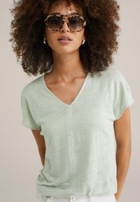 T-shirt vert clair à manches courtes et col en V, fabriqué en tissu doux et texturé. Modèle portant des lunettes de soleil oversize avec une monture écaille de tortue.
