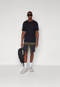 Fekete póló, olíva- és fekete színű zsebes rövidnadrág, fehér csíkos zokni, szürke sportcipő, fekete hátizsákot visel.