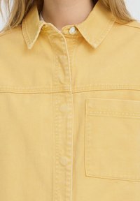 Gros plan sur une chemise en velours côtelé jaune avec col, boutons-pression visibles et poche poitrine portée par une personne aux longs cheveux clairs.