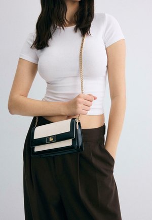 Donna con t-shirt corta bianca e pantaloni scuri che tiene una piccola borsa a tracolla nera e beige con catenella dorata su sfondo neutro.