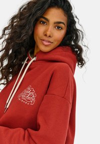 Červená mikina s kapucňou z mäkkého materiálu, s predným logom s nápisom "Radšej by som sa pozeral," obsahuje šnúrku na kapucni a voľný strih.