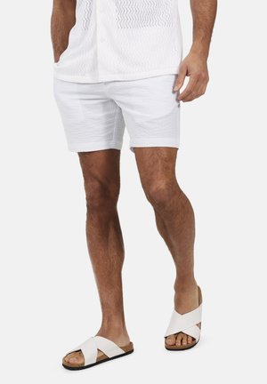 Homme portant un short blanc texturé, une chemise en maille blanche avec les mains dans les poches, et des sandales blanches à brides croisées sur pieds nus.