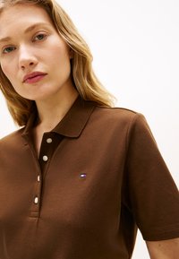 Γυναίκα με ανοιχτόχρωμο δέρμα και ξανθά μαλλιά που φοράει καφέ κοντομάνικο πόλο με μικρό κόκκινο, λευκό και μπλε λογότυπο στο στήθος.