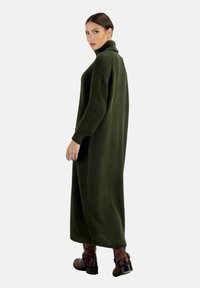 Langes, übergroßes Turtleneck-Strickkleid in Dunkelgrün, aus strukturiertem Strickstoff. Mit langen Ärmeln und seitlichen Schlitzen. Kombiniert mit braunen Schnallenstiefeln.