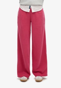 Pantaloni della tuta larghi rosa realizzati in morbido tessuto, con un elastico in vita e un sottile dettaglio del logo. Abbinati a una maglietta bianca e scarpe da ginnastica.