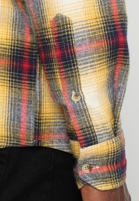 Camicia a quadri gialla realizzata in tessuto con linee rosse e blu. Presenta polsini con bottoni e una texture spazzolata per una maggiore morbidezza.