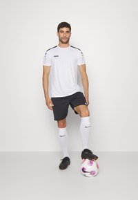 Maglietta sportiva bianca con accenti neri e logo, pantaloni corti neri, calzini bianchi, scarpe da calcio nere e un pallone da calcio multicolore ai suoi piedi.