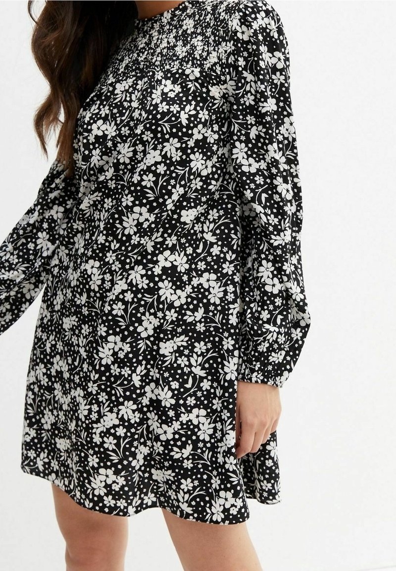 Robe noire ornée d'un motif floral blanc, manches longues et encolure smockée. Le tissu semble léger avec une silhouette fluide.