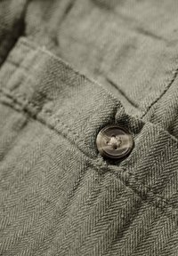 Olijfgroene stof met een visgraat patroon; heeft een ronde, bruine knop, stiksel details en een gestructureerd oppervlak.
