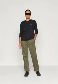 Mulher aStanding com uma mão no bolso, vestindo uma camisa preta de mangas compridas, calças verde-oliva, sapatos pretos e óculos de sol oversized pretos.