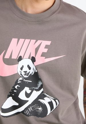 Graues T-Shirt mit pinkem Nike-Logo und Grafik eines Pandas, der auf einem schwarz-weißen Nike-Sneaker sitzt.