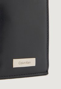 Jūras zila ādas maku, kuram ir sudraba taisnstūra zīmola plāksne ar gravējumu "Calvin Klein" un kontrastējoša šuvēšana malu garumā.