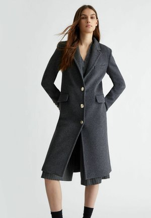 Cappotto di lana grigio con colletto sciallato, design a un solo petto, bottoni dorati e tasche a patta. Presenta un taglio lungo fino al ginocchio e un tessuto texture.