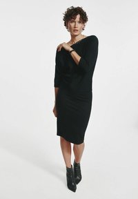 Kobieta z krótkimi, kręconymi włosami, ubrana w czarną bluzkę z rękawami 3/4, pasującą spódnicę do kolan oraz czarne botki, pozująca na tle białym.