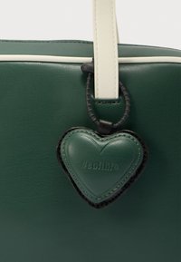Πράσινη τσάντα tote από δέρμα με άσπρο περίγραμμα και υφή στην επιφάνεια. Διαθέτει κρεμαστό κόσμημα σε σχήμα καρδιάς με την επιγραφή "#softlife" προσαρτημένο σε μαύρο λουράκι.