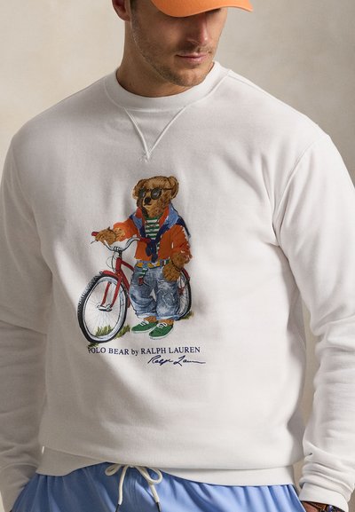 Egy férfi fehér pulóvert visel, amin napszemüveges medve grafikája látható, narancssárga dzsekit, farmert és zöld cipőt visel, miközben egy kerékpár mellett áll.