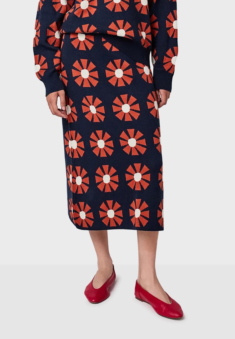 Mornarskoplava suknja i set s puloverom s uzorkom crvenih i bijelih cvjetova, nosi se s crvenim baletankama na svijetloj pozadini.