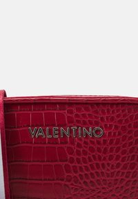 Červená kožená kabelka s krokodýlím vzorem a kovovým logem "VALENTINO". Disponuje elegantním designem a texturovaným povrchem.
